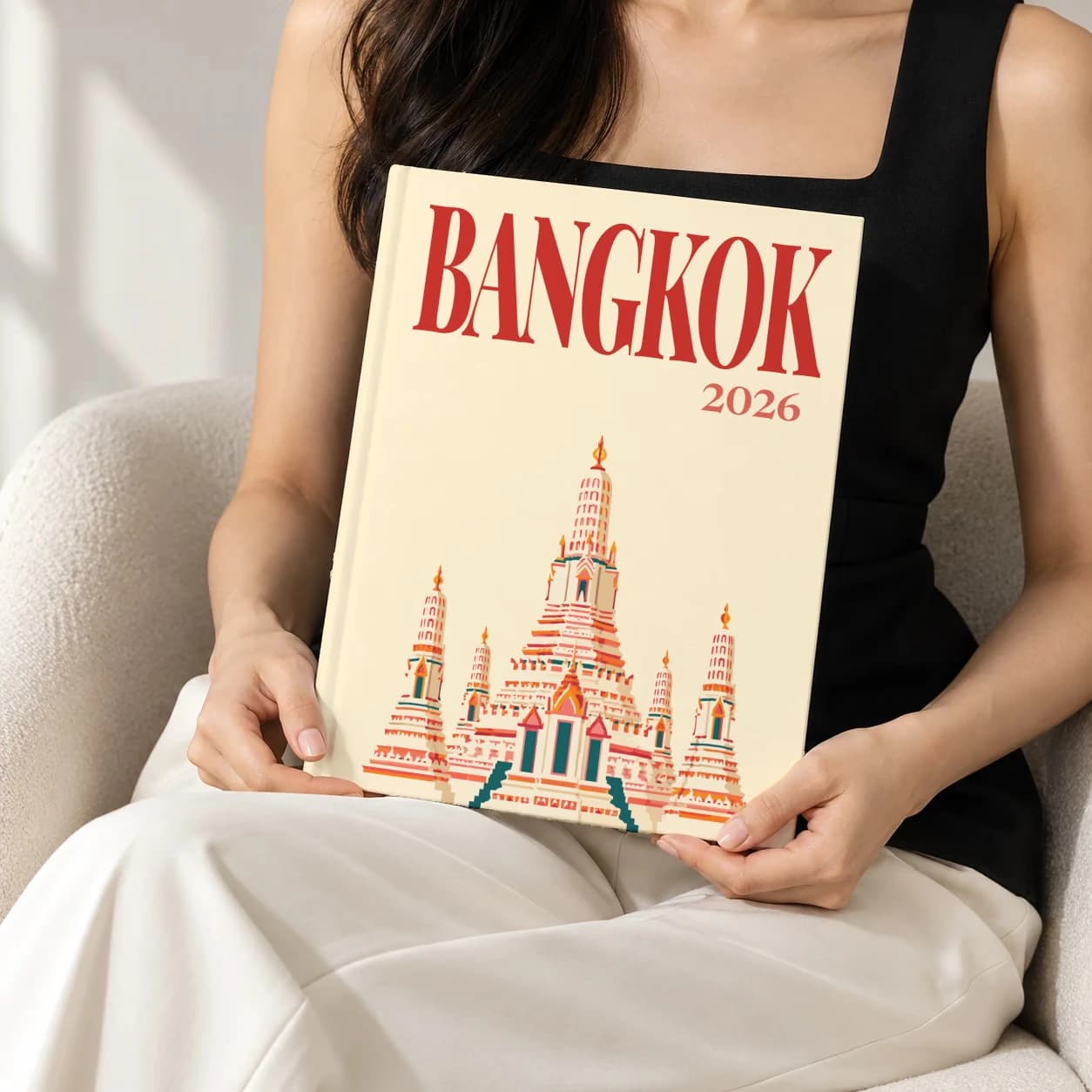 Bangkok hardcover photo book with the illustrated cover of the Wat Arun, held upright in two hands by a woman in a black top, sitting in a cream boucle armchair