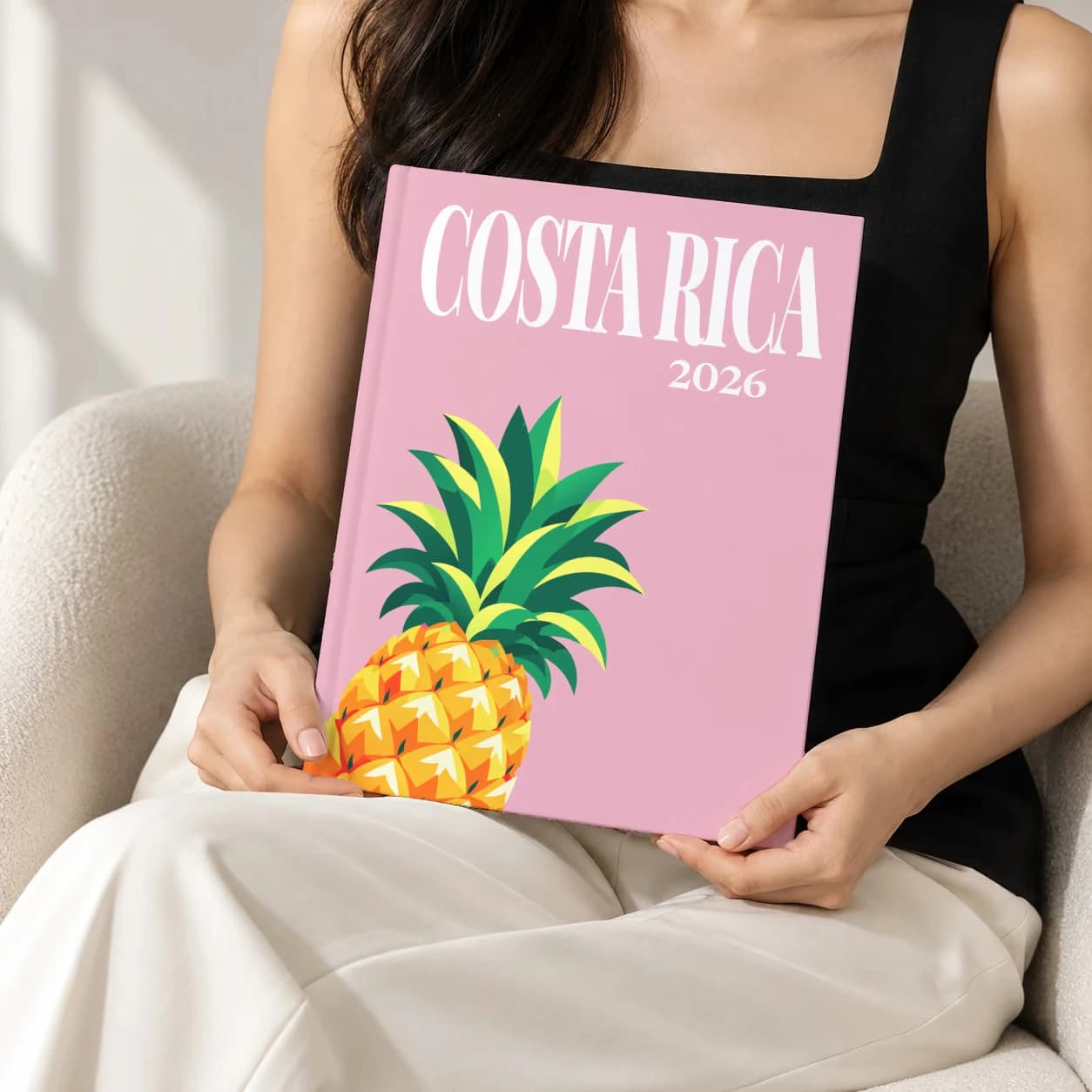 Costa Rica hardcover photo book with the illustrated cover of the pineapple, held upright in two hands by a woman in a black top, sitting in a cream boucle armchair