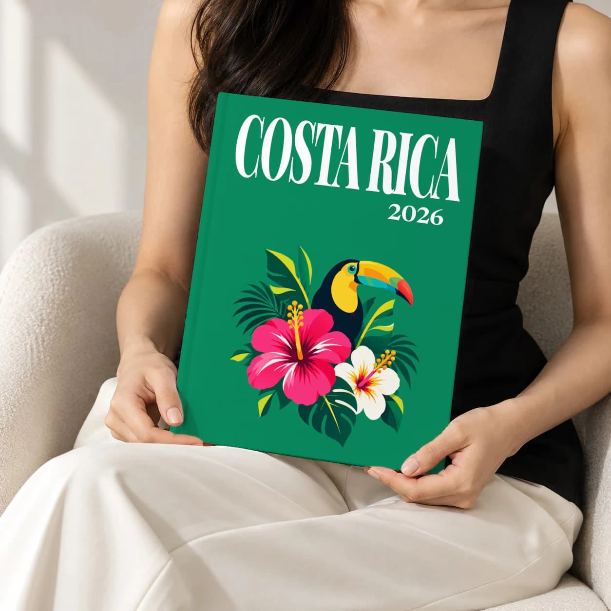 Costa Rica hardcover photo book with the illustrated cover of the toucan and hibiscus, held upright in two hands by a woman in a black top, sitting in a cream boucle armchair