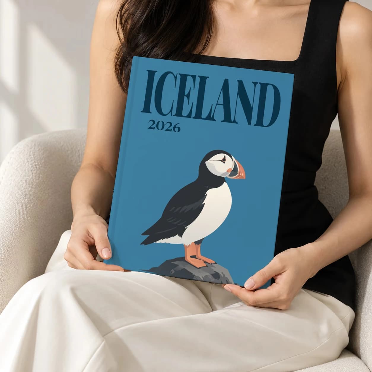 Iceland hardcover photo book with the illustrated cover of the puffin, held upright in two hands by a woman in a black top, sitting in a cream boucle armchair