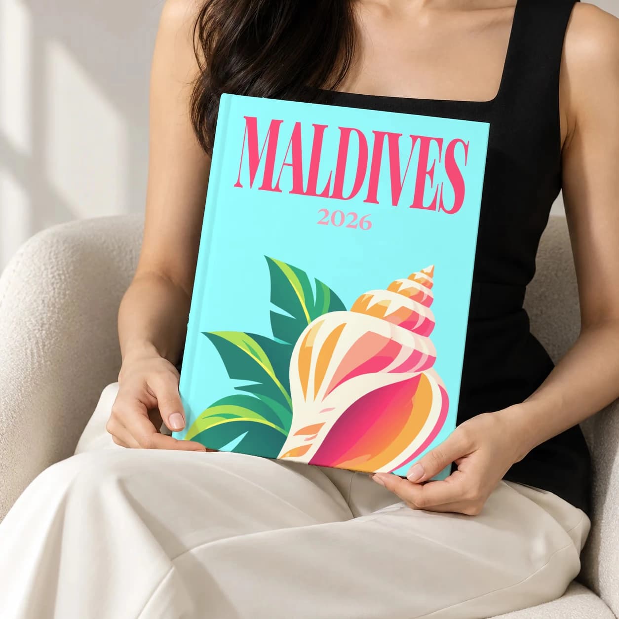 Maldives hardcover photo book with the illustrated cover of the conch shell, held upright in two hands by a woman in a black top, sitting in a cream boucle armchair