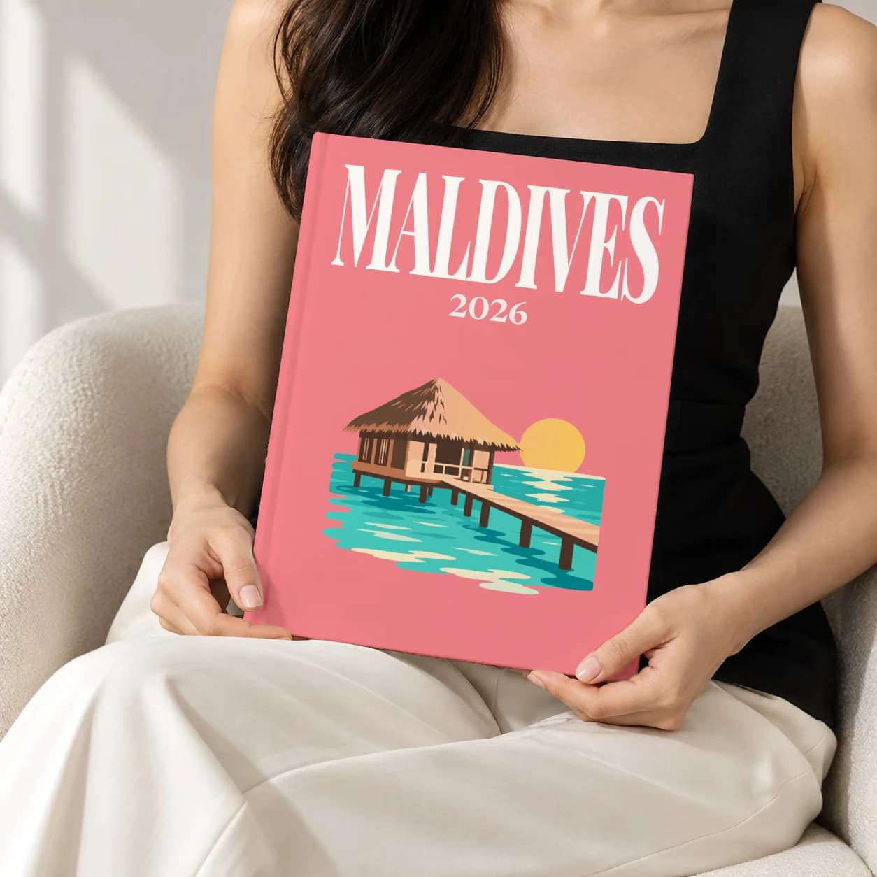 Maldives hardcover photo book with the illustrated cover of the water villa, held upright in two hands by a woman in a black top, sitting in a cream boucle armchair