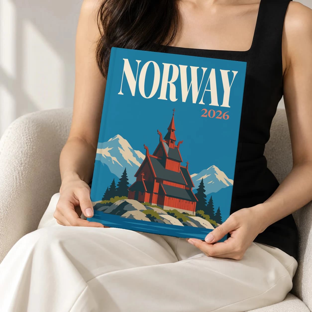 Norway hardcover photo book with the illustrated cover of the stave church, held upright in two hands by a woman in a black top, sitting in a cream boucle armchair