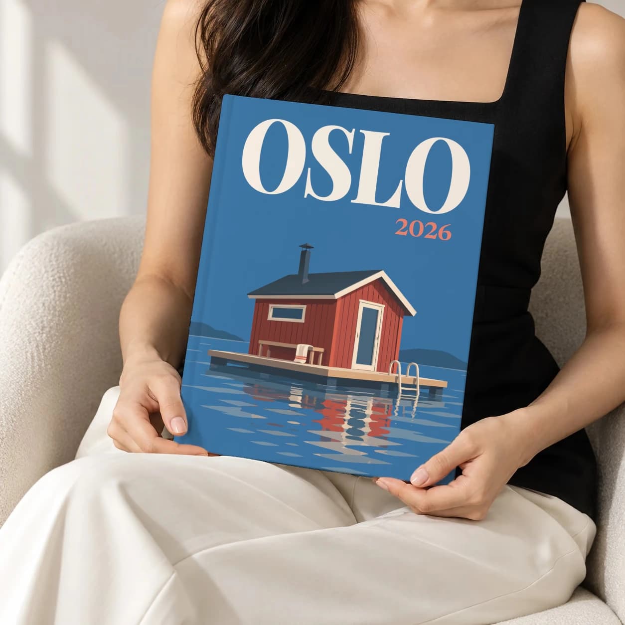 Oslo hardcover photo book with the illustrated cover of the floating sauna, held upright in two hands by a woman in a black top, sitting in a cream boucle armchair