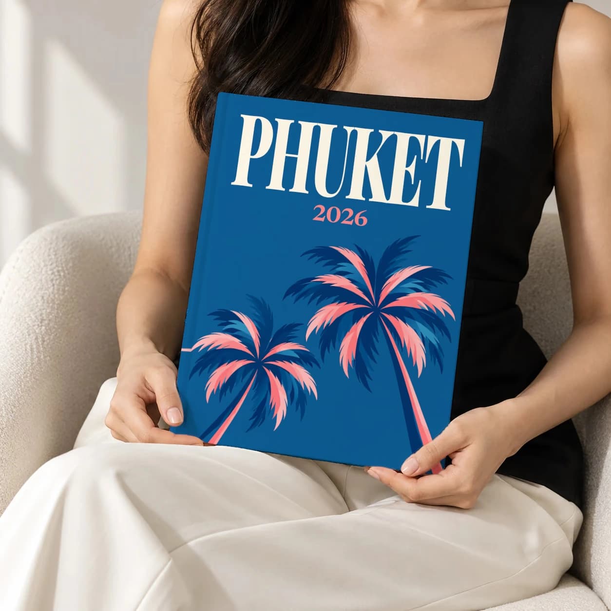 Phuket hardcover photo book with the illustrated cover of the palms, held upright in two hands by a woman in a black top, sitting in a cream boucle armchair