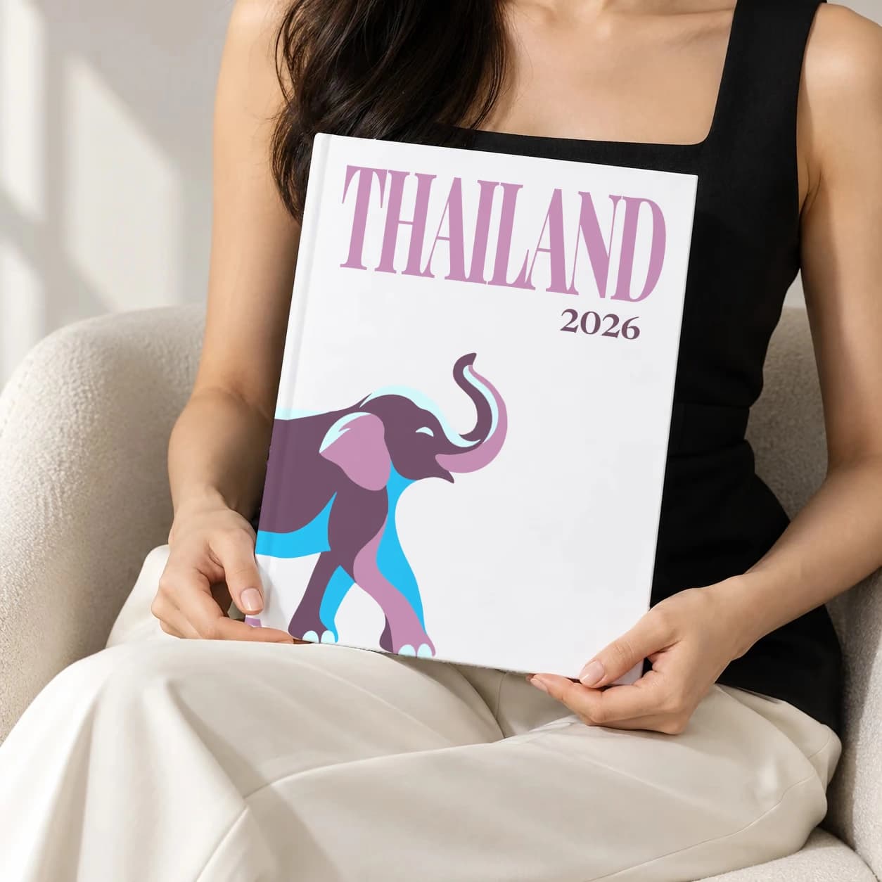 Thailand hardcover photo book with the illustrated cover of an elephant on white, held upright in two hands by a woman in a black top, sitting in a cream boucle armchair