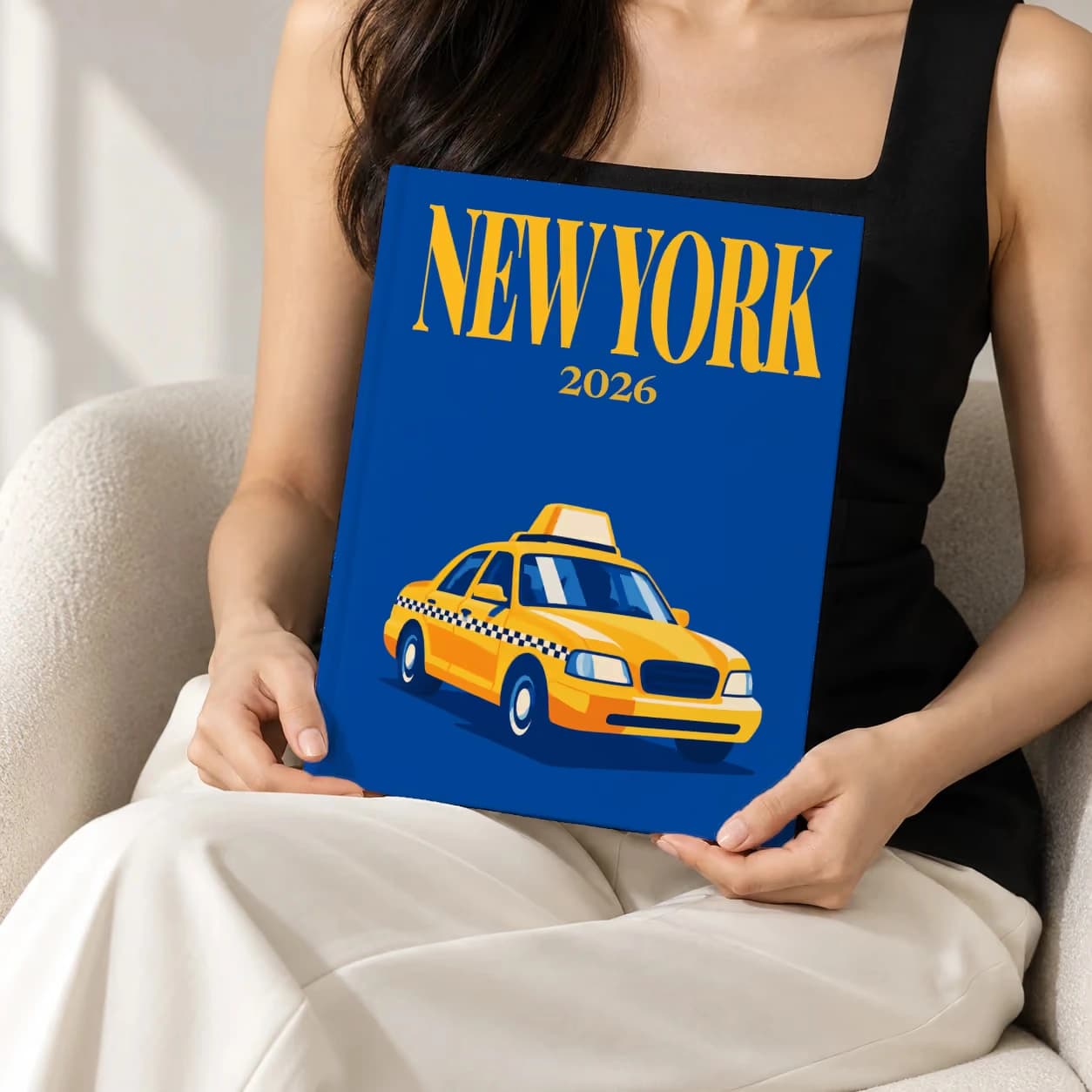 New York hardcover photo book with an illustrated cover of a yellow taxi on royal blue, held upright in two hands by a woman in a black top, sitting in a cream boucle armchair