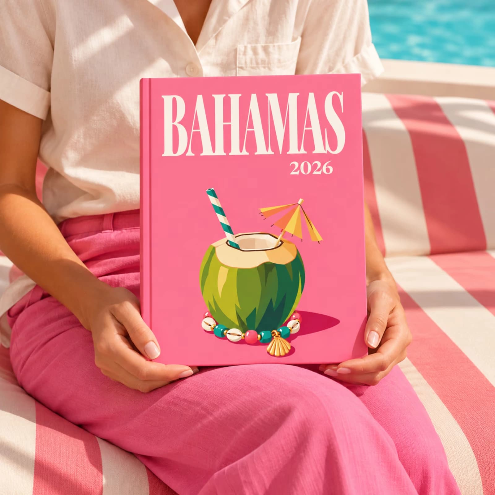 A woman on a pink striped sofa holding a Bahamas travel photo book with an illustrated coconut drink