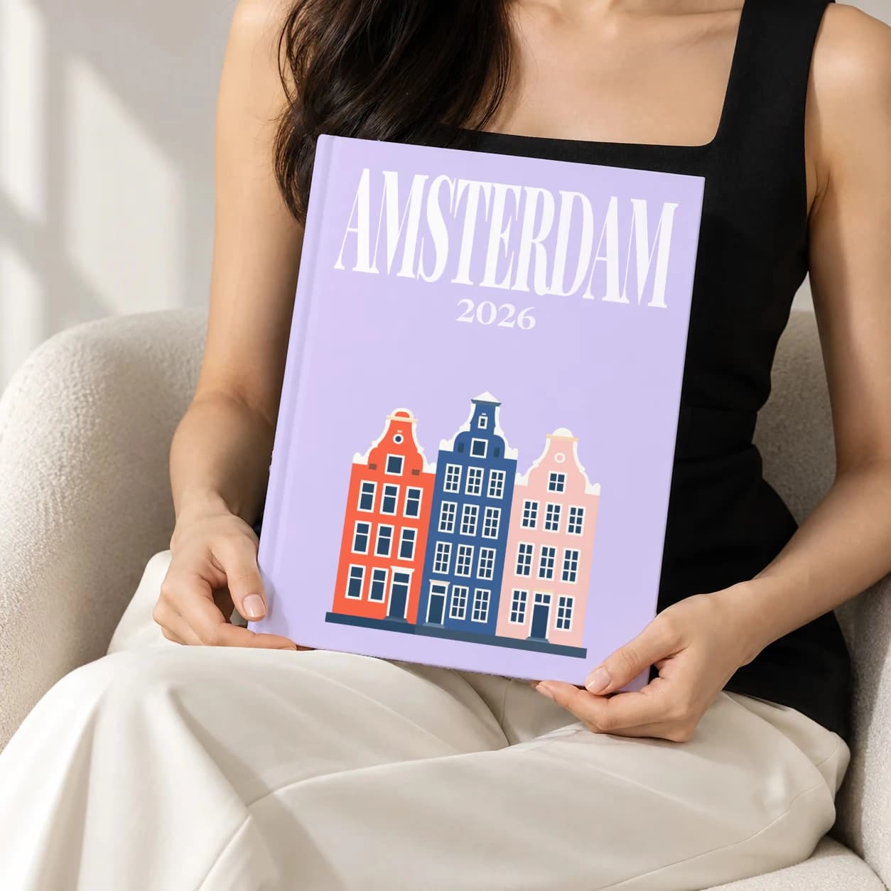 Amsterdam hardcover photo book with the illustrated cover of the canal houses, held upright in two hands by a woman in a black top, sitting in a cream boucle armchair
