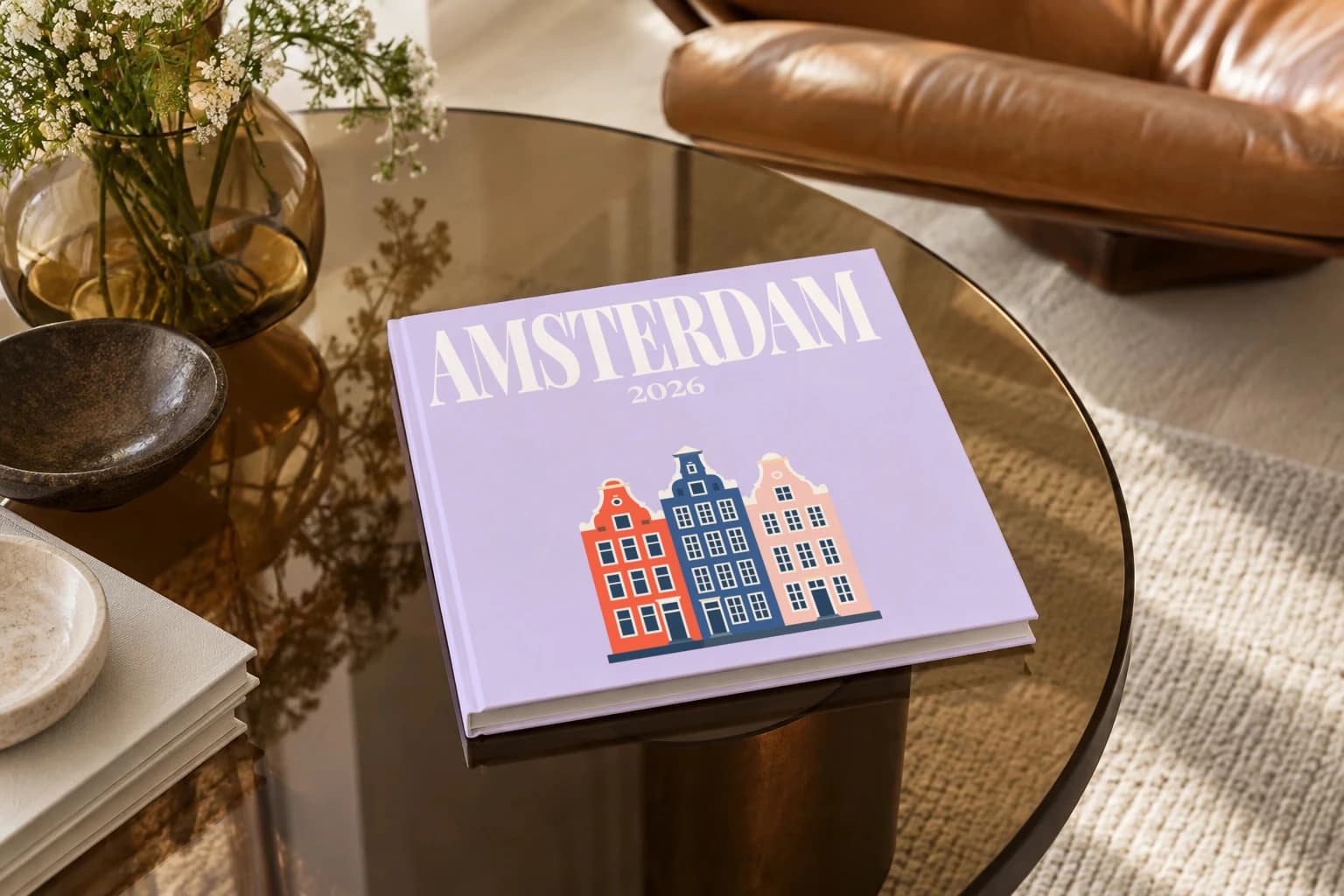 Amsterdam square hardcover photo book with the illustrated cover of the canal houses, on a smoked glass coffee table beside a leather armchair in warm afternoon light
