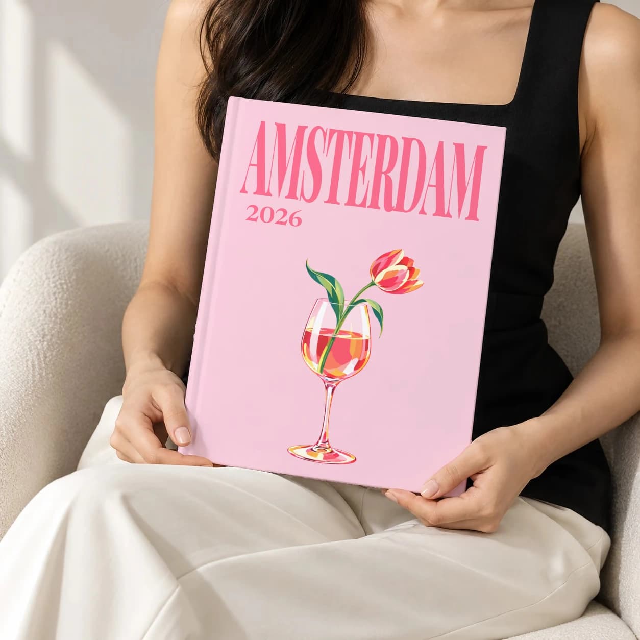 Amsterdam hardcover photo book with the illustrated cover of the tulip in wine glass, held upright in two hands by a woman in a black top, sitting in a cream boucle armchair
