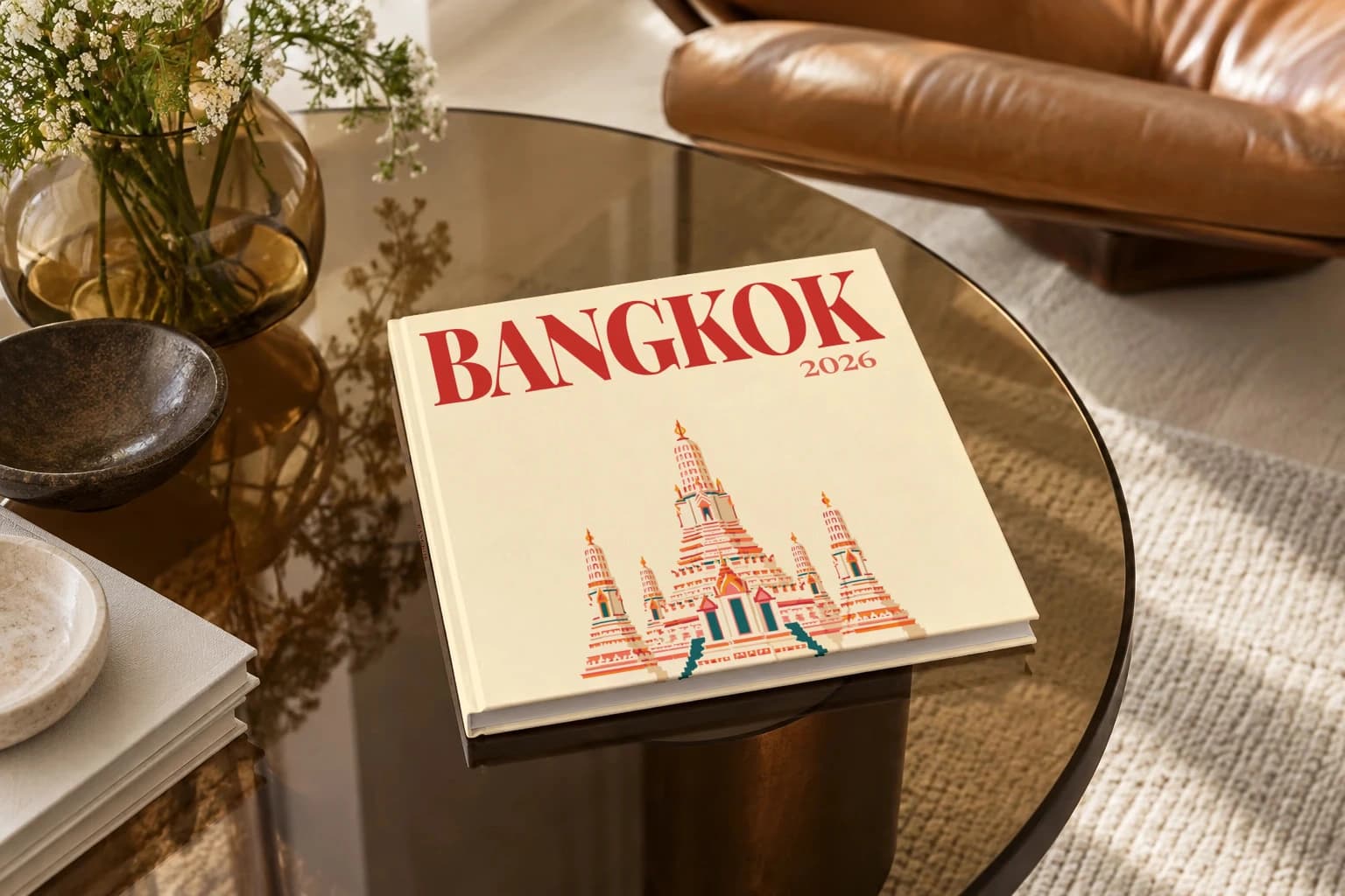 Bangkok square hardcover photo book with the illustrated cover of the Wat Arun, on a smoked glass coffee table beside a leather armchair in warm afternoon light