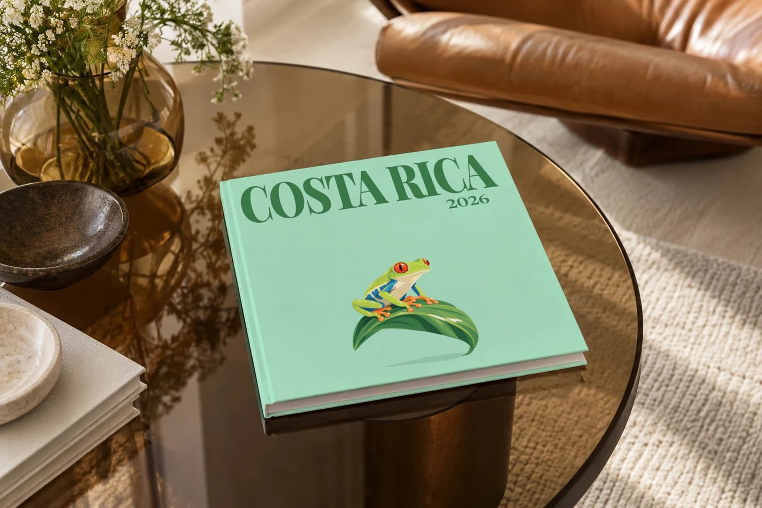 Costa Rica square hardcover photo book with the illustrated cover of the red-eyed tree frog, on a smoked glass coffee table beside a leather armchair in warm afternoon light