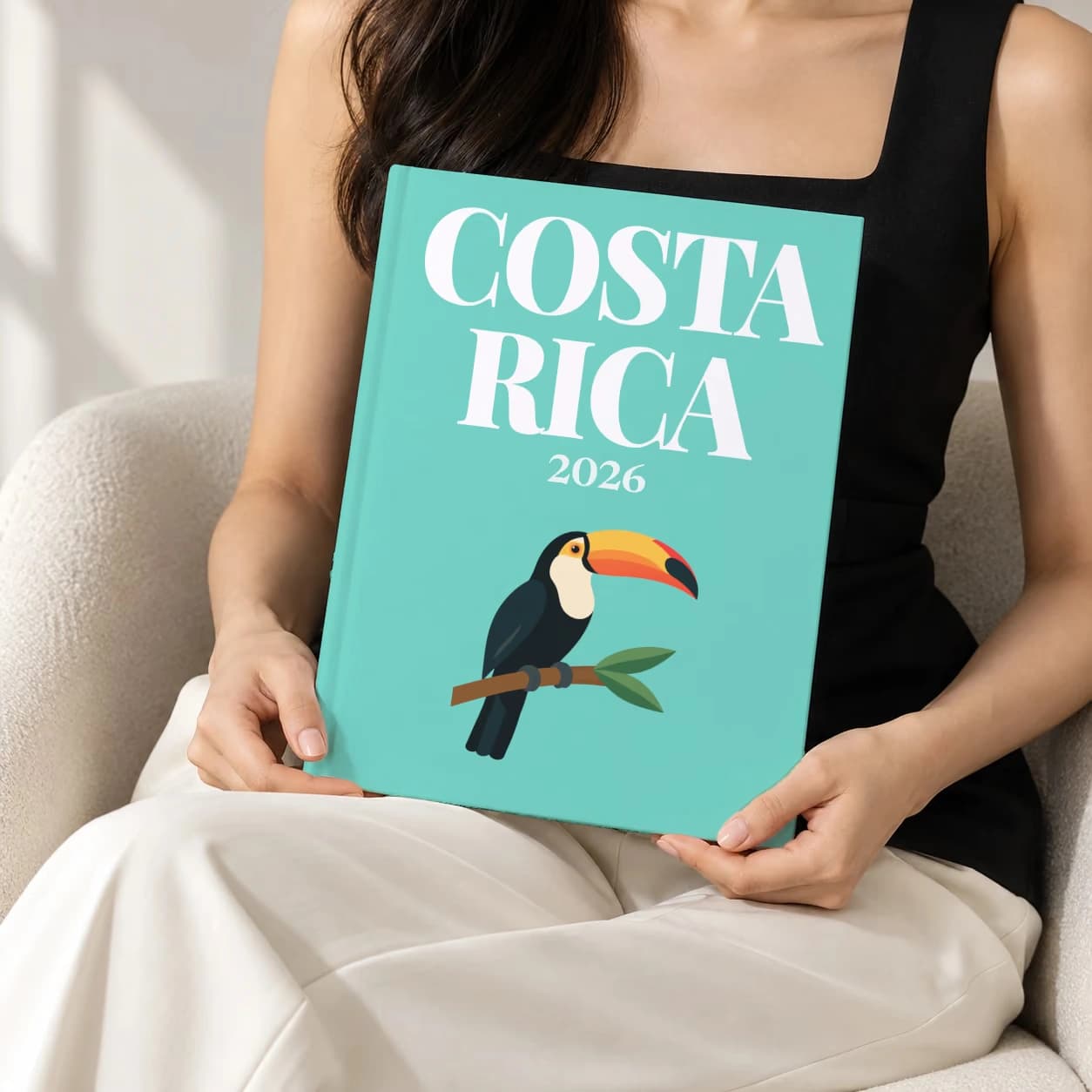 Costa Rica hardcover photo book with the illustrated cover of the toucan on a branch, held upright in two hands by a woman in a black top, sitting in a cream boucle armchair