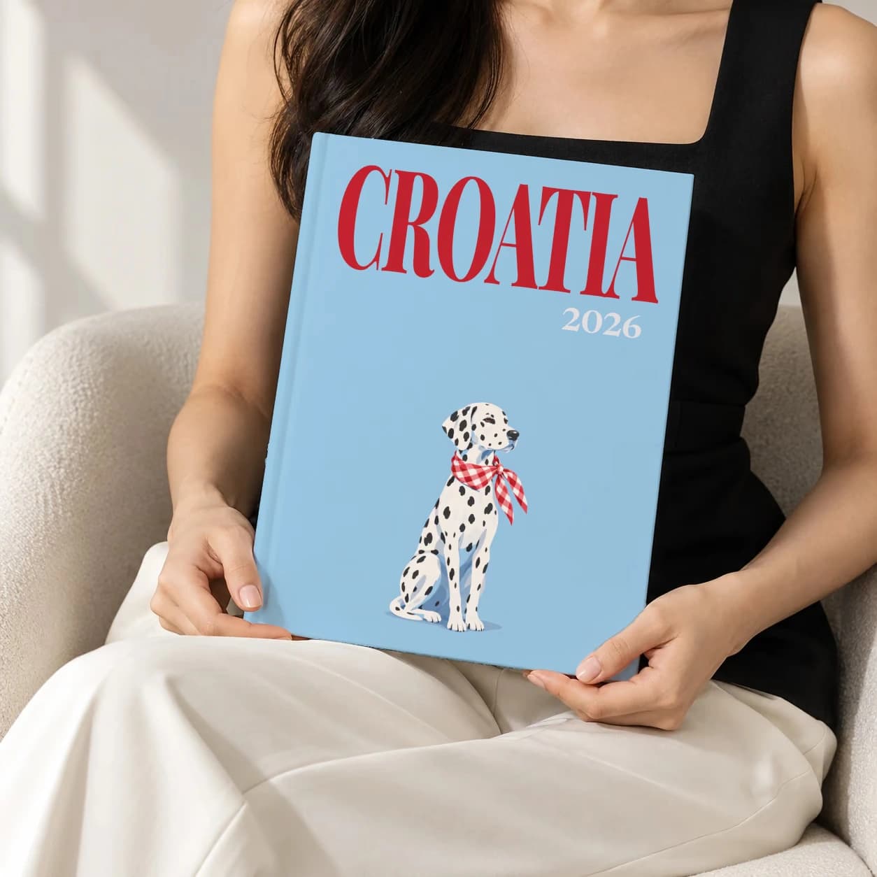 Croatia hardcover photo book with the illustrated cover of the dalmatian, held upright in two hands by a woman in a black top, sitting in a cream boucle armchair