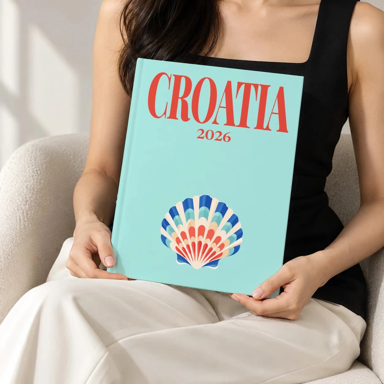 Croatia hardcover photo book with the illustrated cover of the shell, held upright in two hands by a woman in a black top, sitting in a cream boucle armchair