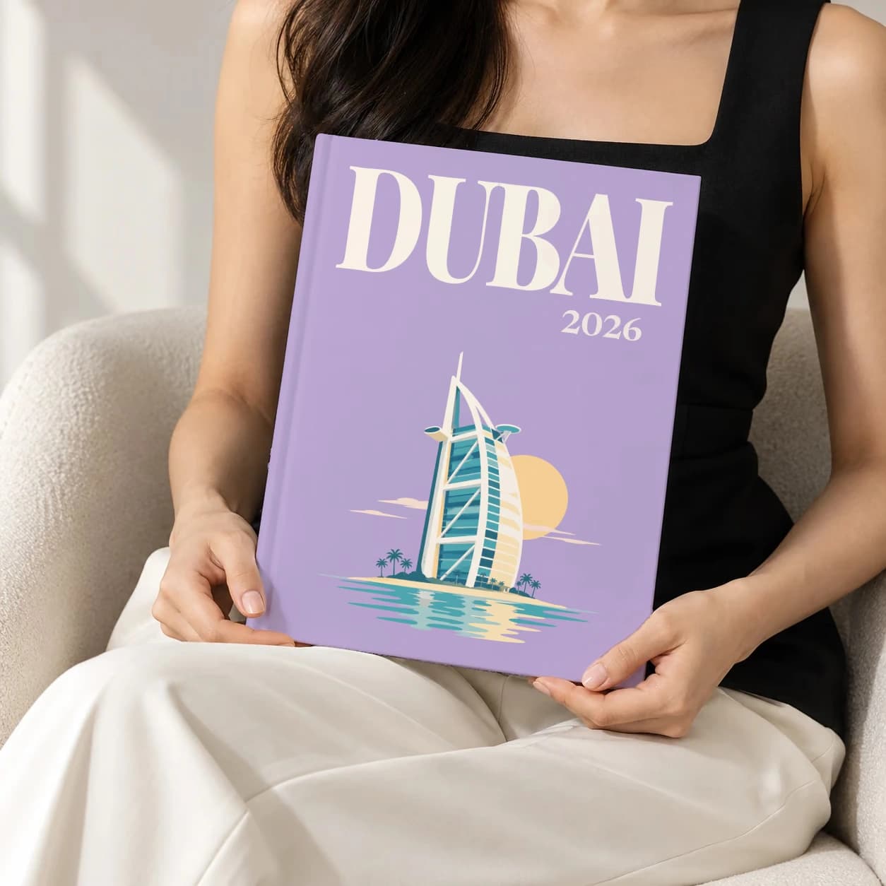 Dubai hardcover photo book with the illustrated cover of the Burj Al Arab artwork in lavender, held upright in two hands by a woman in a black top, sitting in a cream boucle armchair