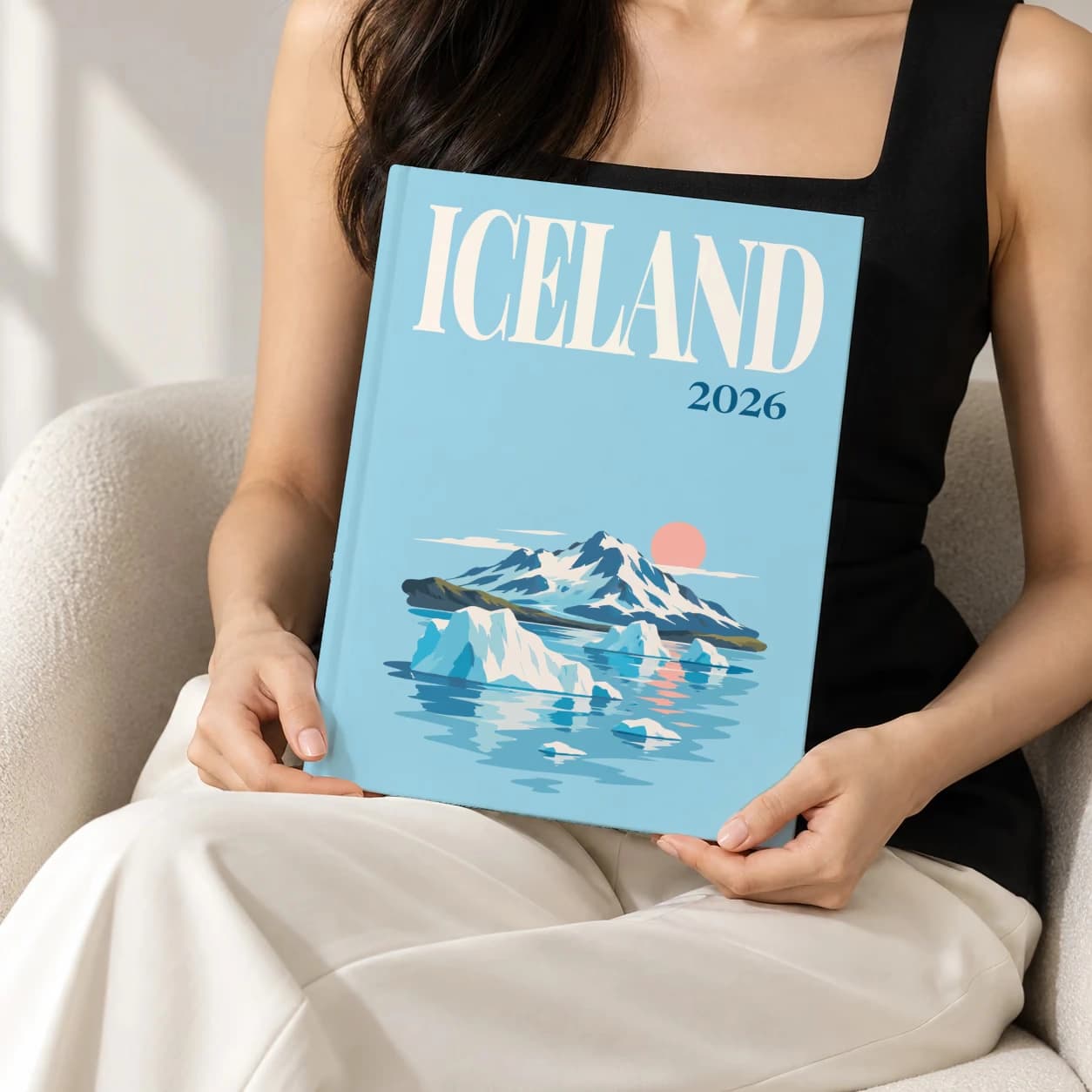Iceland hardcover photo book with the illustrated cover of the icebergs at sunset, held upright in two hands by a woman in a black top, sitting in a cream boucle armchair