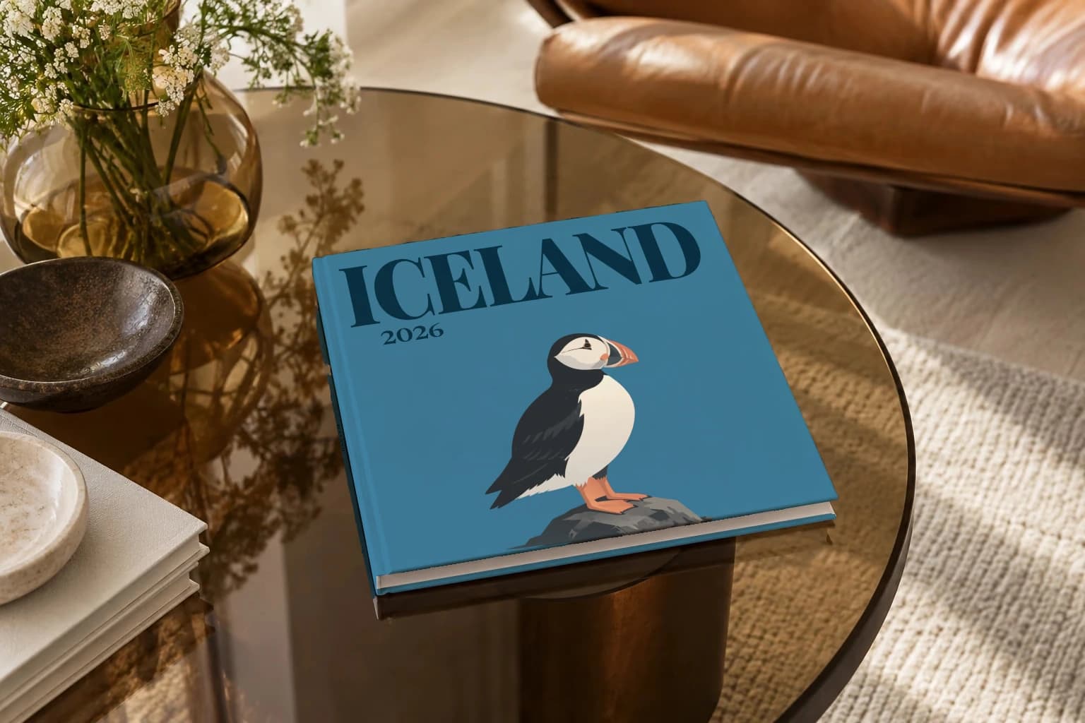 Iceland square hardcover photo book with the illustrated cover of the puffin, on a smoked glass coffee table beside a leather armchair in warm afternoon light