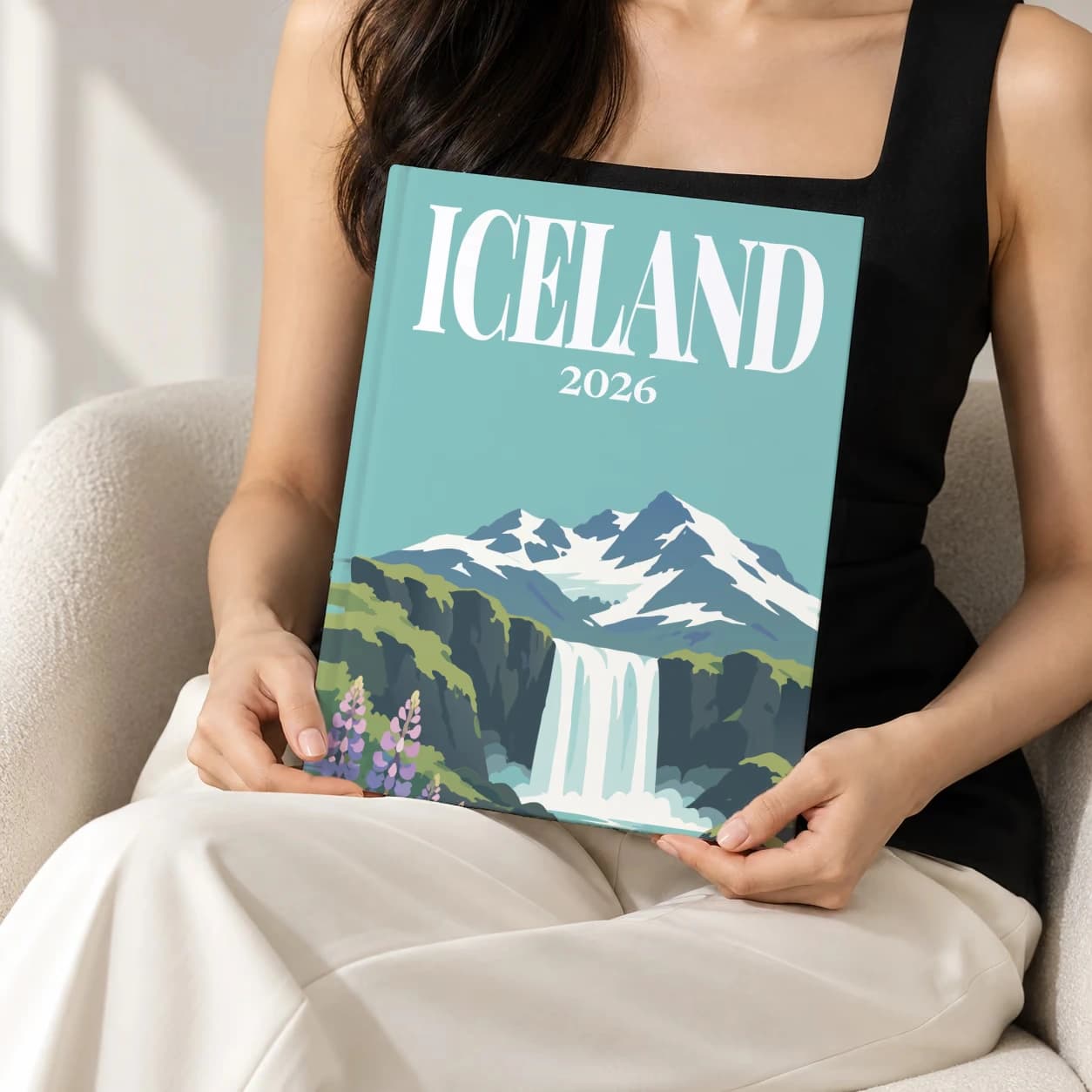 Iceland hardcover photo book with the illustrated cover of the waterfall and lupins, held upright in two hands by a woman in a black top, sitting in a cream boucle armchair