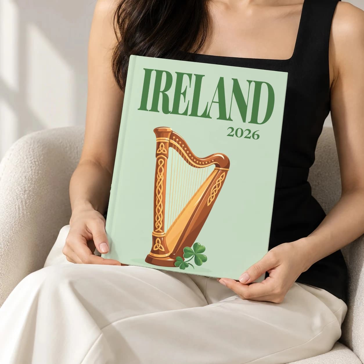 Ireland hardcover photo book with the illustrated cover of the Celtic harp, held upright in two hands by a woman in a black top, sitting in a cream boucle armchair