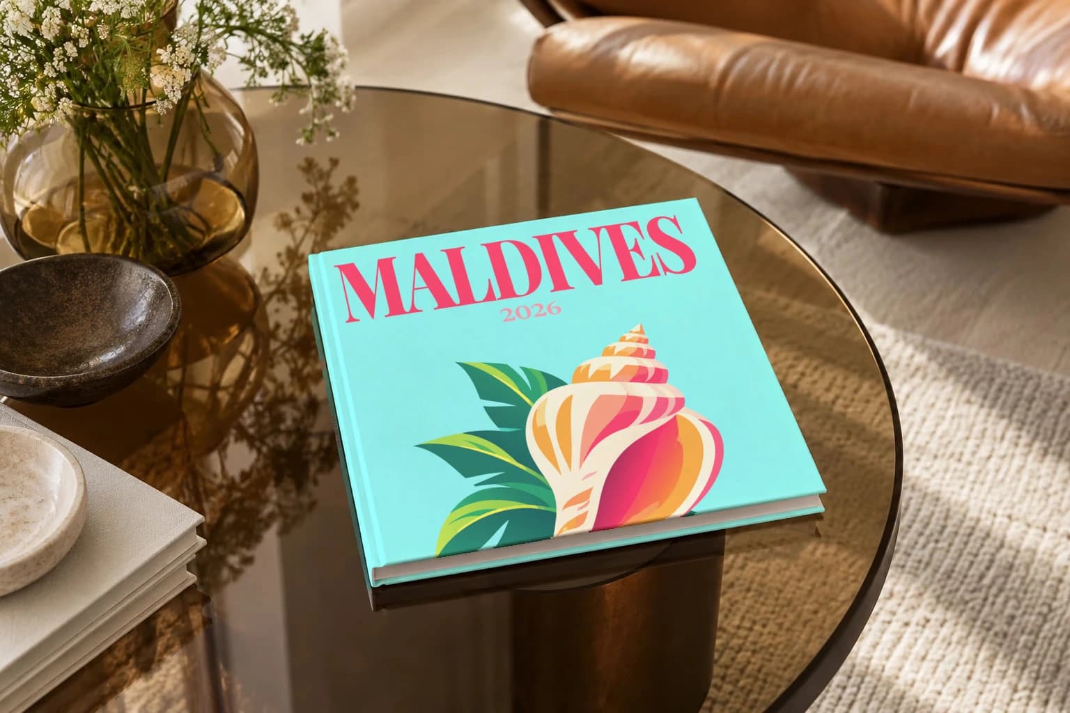 Maldives square hardcover photo book with the illustrated cover of the conch shell, on a smoked glass coffee table beside a leather armchair in warm afternoon light