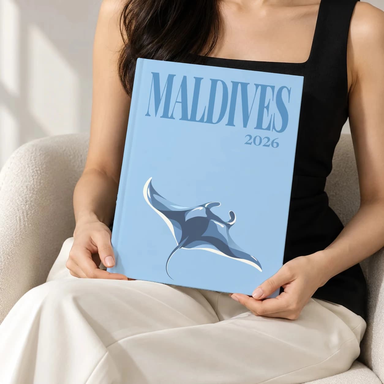 Maldives hardcover photo book with the illustrated cover of the manta ray, held upright in two hands by a woman in a black top, sitting in a cream boucle armchair