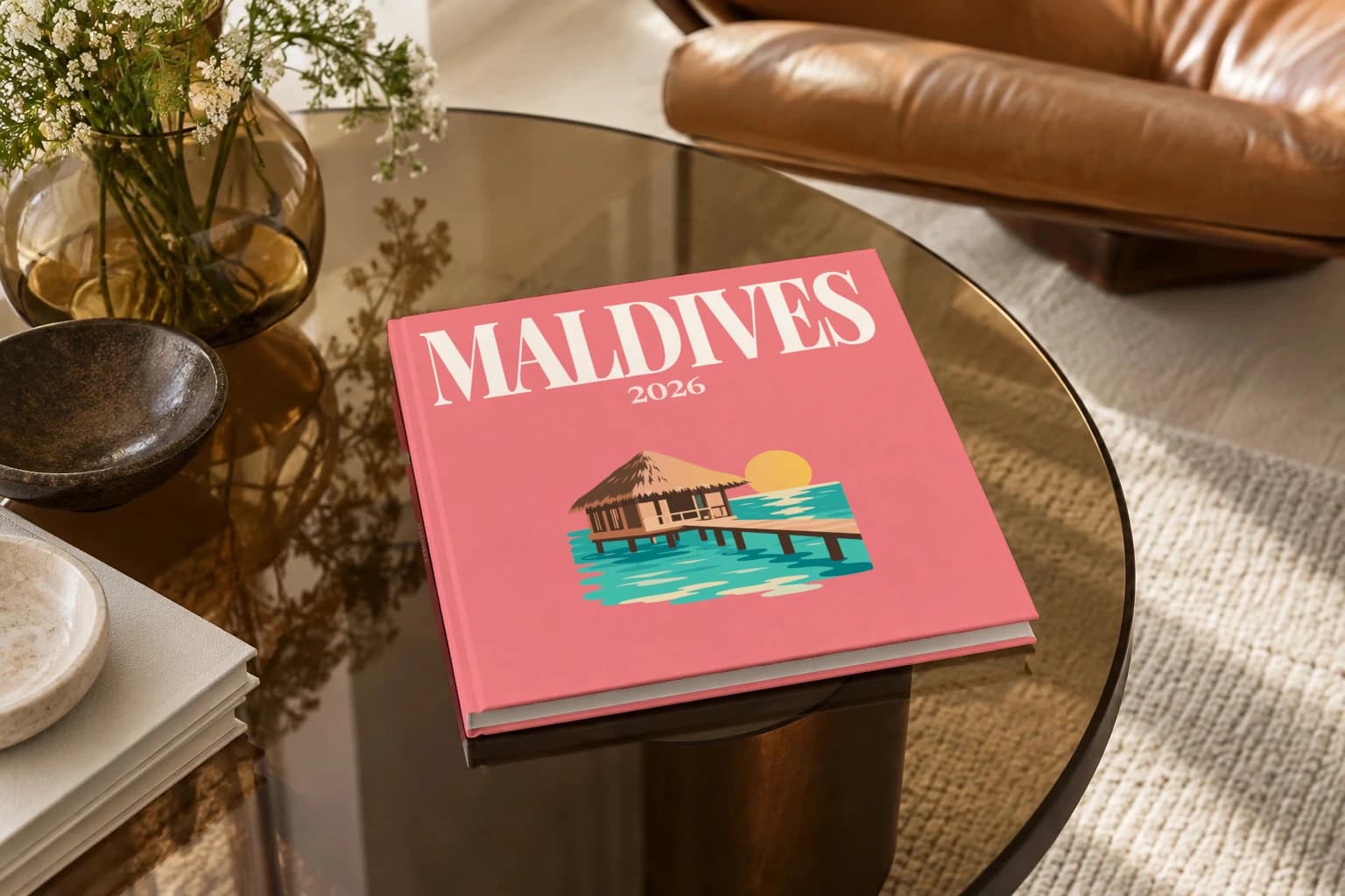 Maldives square hardcover photo book with the illustrated cover of the water villa, on a smoked glass coffee table beside a leather armchair in warm afternoon light