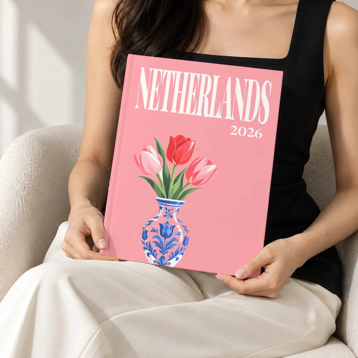 Netherlands hardcover photo book with the illustrated cover of the tulips, held upright in two hands by a woman in a black top, sitting in a cream boucle armchair