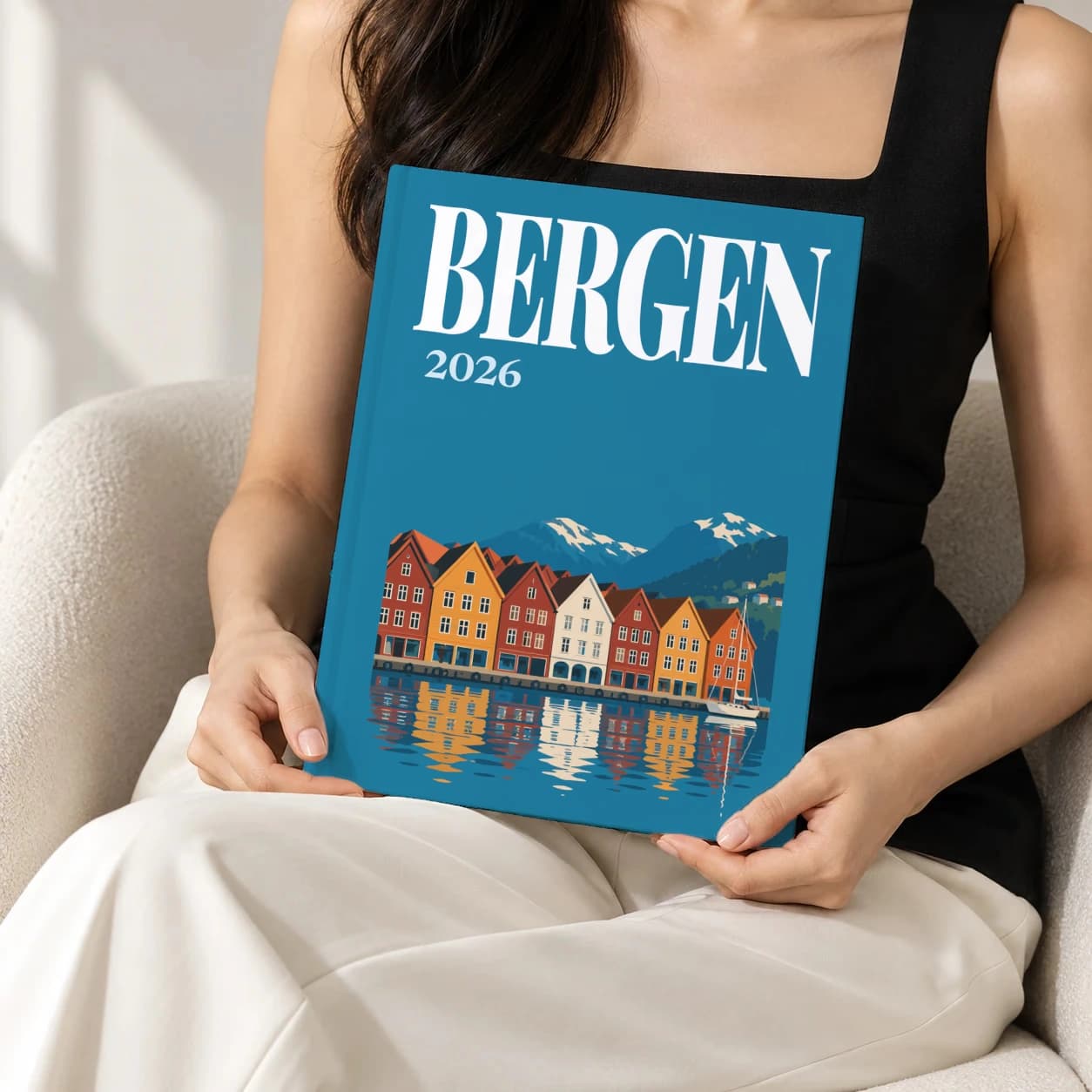 Bergen hardcover photo book with the illustrated cover of the Bryggen wharf, held upright in two hands by a woman in a black top, sitting in a cream boucle armchair