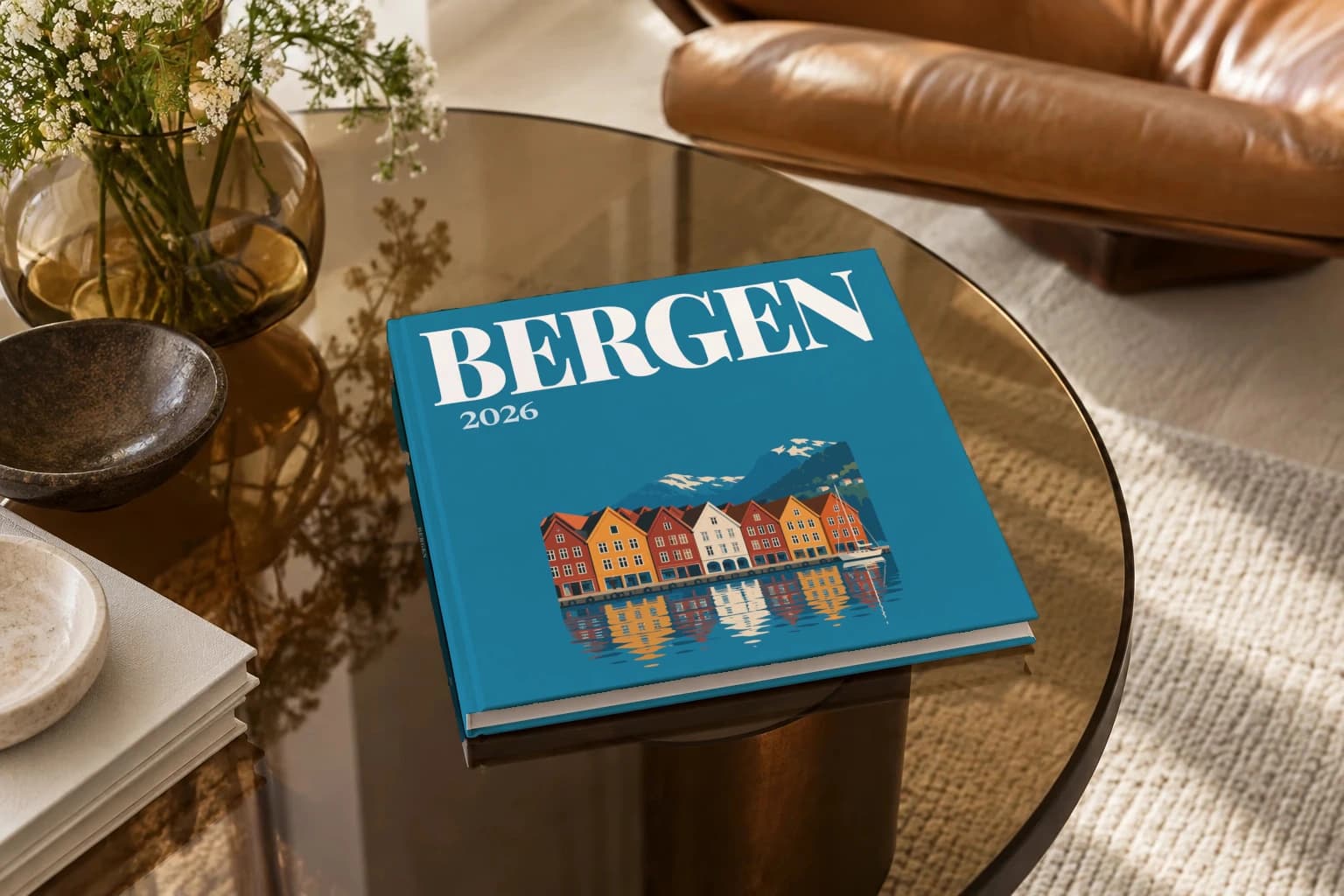 Bergen square hardcover photo book with the illustrated cover of the Bryggen wharf, on a smoked glass coffee table beside a leather armchair in warm afternoon light
