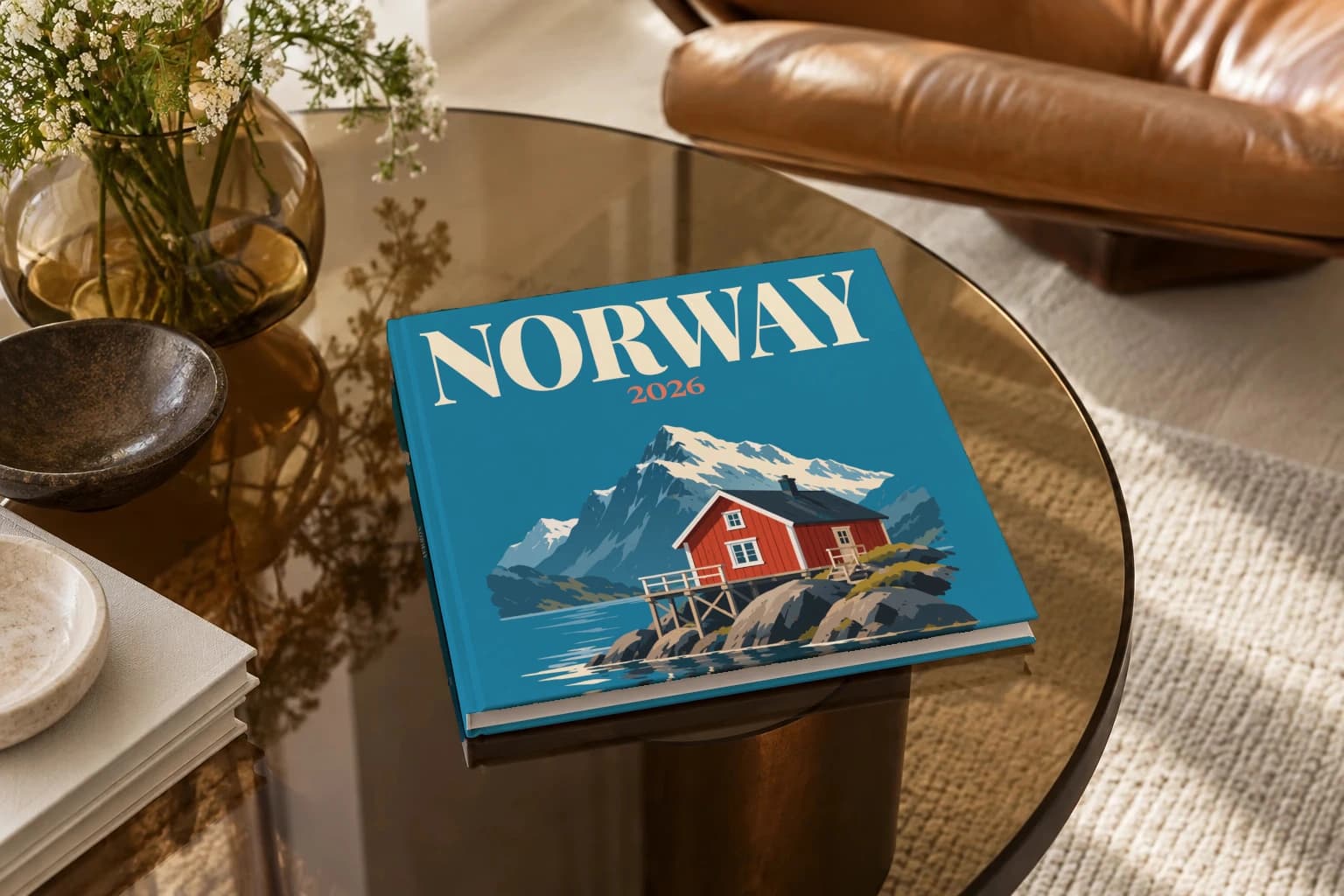 Norway square hardcover photo book with the illustrated cover of the rorbu cabin, on a smoked glass coffee table beside a leather armchair in warm afternoon light