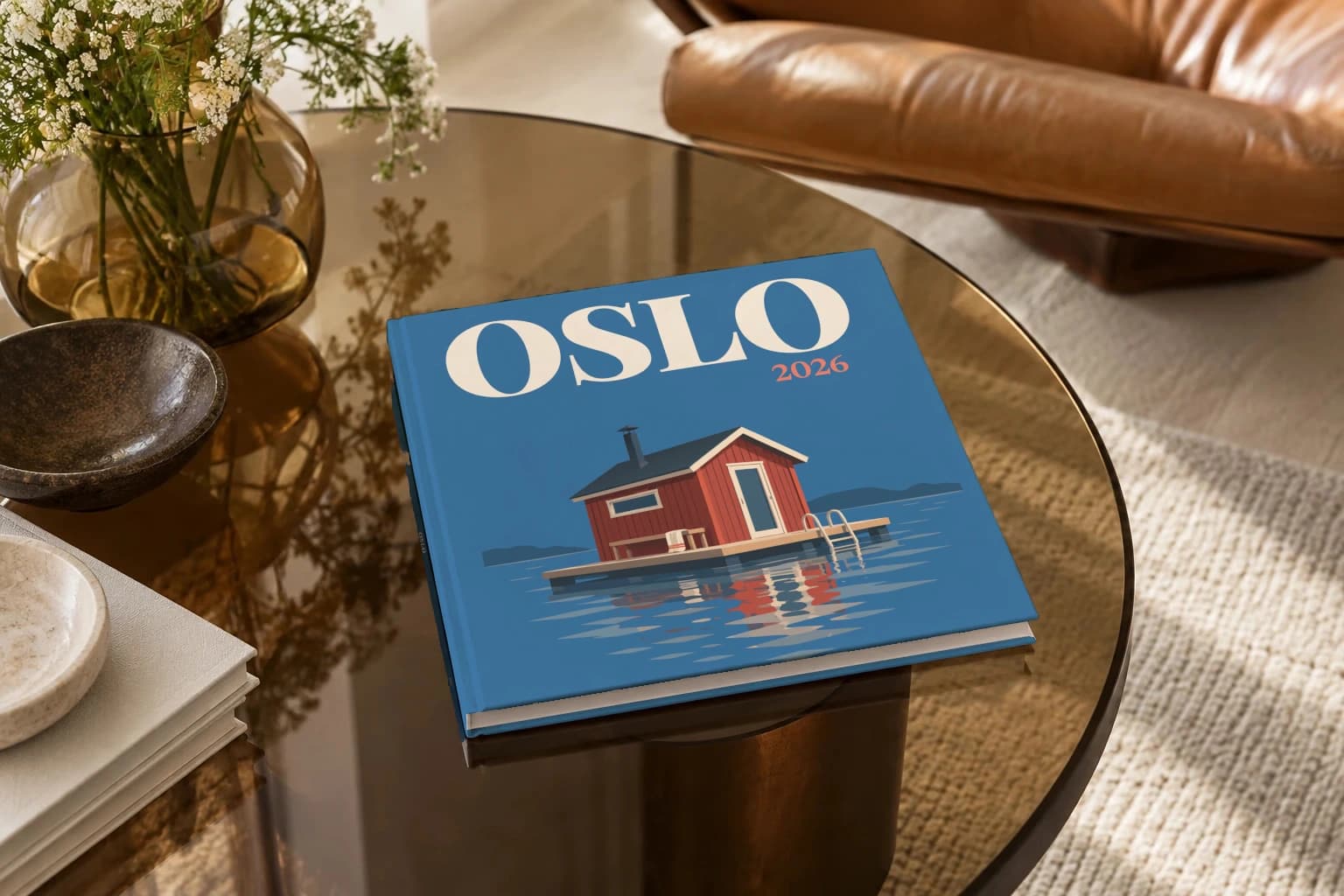 Oslo square hardcover photo book with the illustrated cover of the floating sauna, on a smoked glass coffee table beside a leather armchair in warm afternoon light
