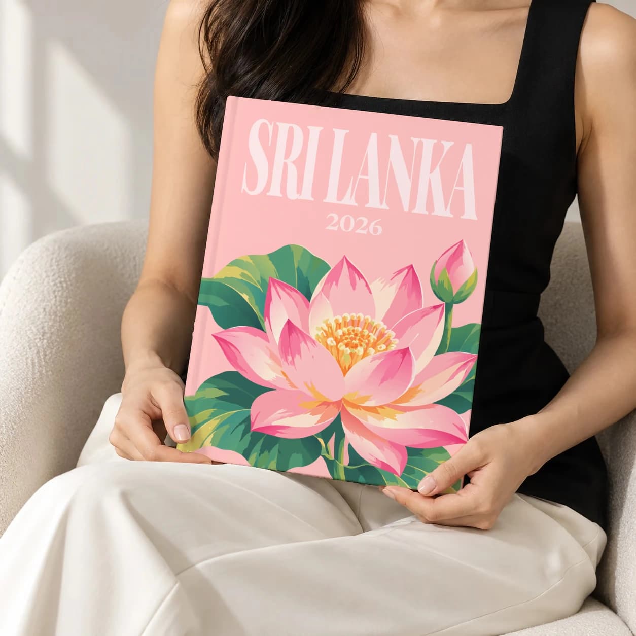 Sri Lanka hardcover photo book with the illustrated cover of the lotus, held upright in two hands by a woman in a black top, sitting in a cream boucle armchair