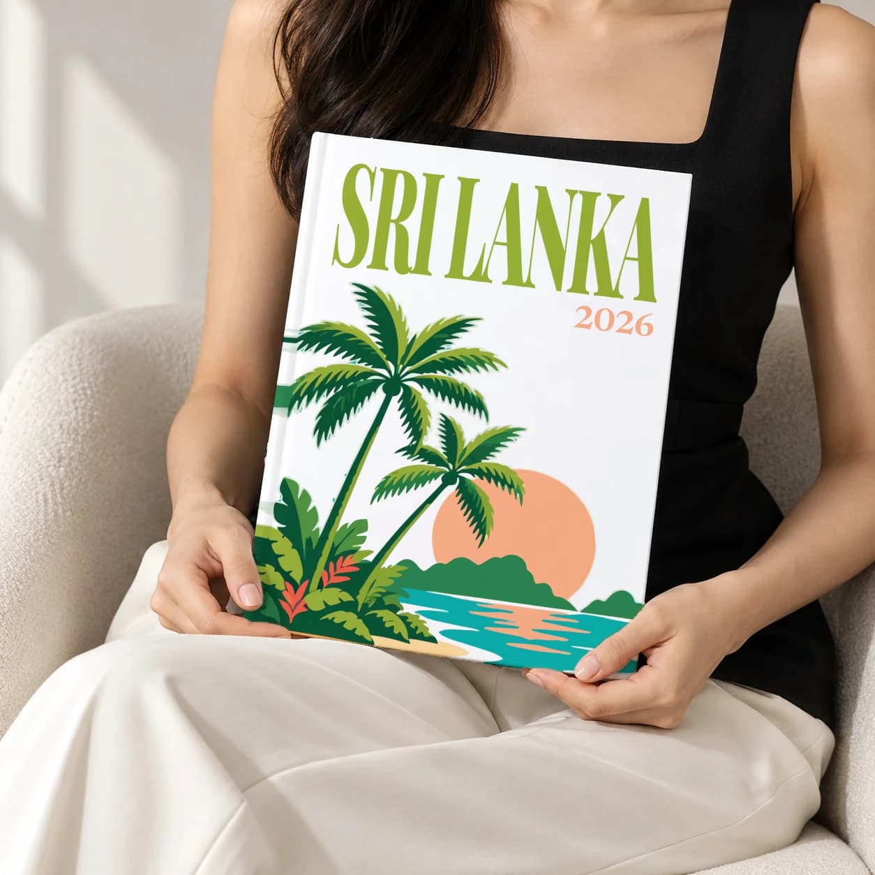 Sri Lanka hardcover photo book with the illustrated cover of the palm beach, held upright in two hands by a woman in a black top, sitting in a cream boucle armchair