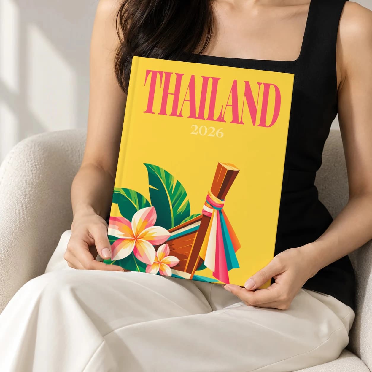 Thailand hardcover photo book with the illustrated cover of the longtail boat, held upright in two hands by a woman in a black top, sitting in a cream boucle armchair