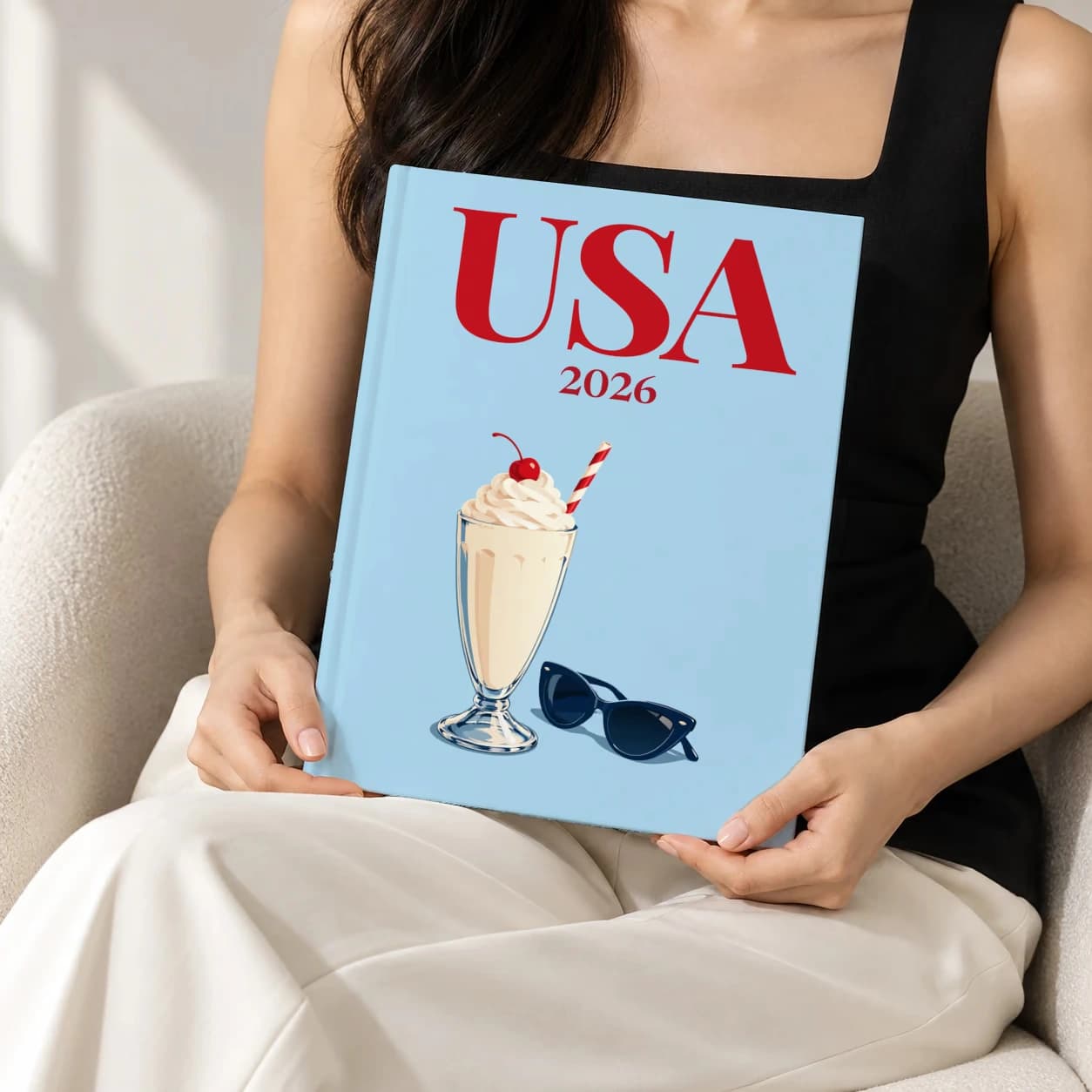 United States hardcover photo book with the USA illustrated cover of the milkshake and sunglasses, held upright in two hands by a woman in a black top, sitting in a cream boucle armchair