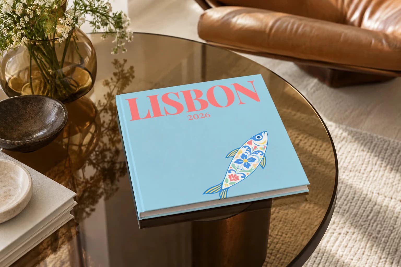 Lisbon hardcover photo book with an illustrated cover of a sardine painted like azulejo tiles, on a smoked glass coffee table beside a leather armchair in warm afternoon light