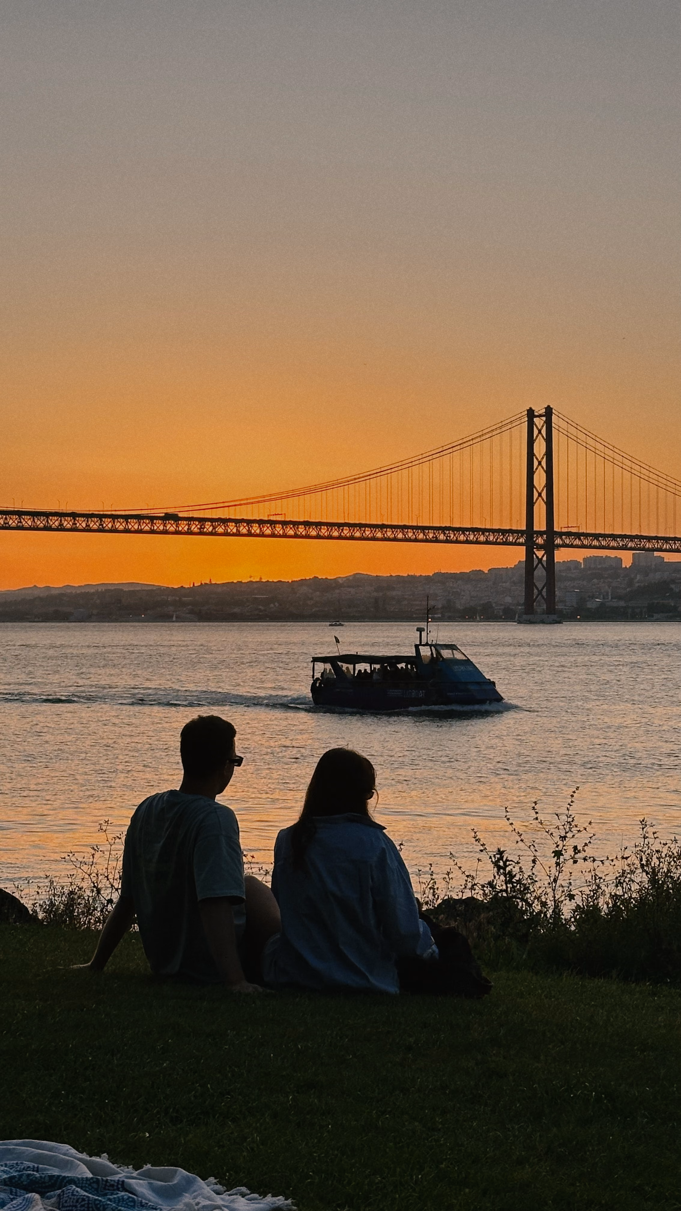 Two people watching a boat pass beneath a suspension bridge at sunset in Lisbon