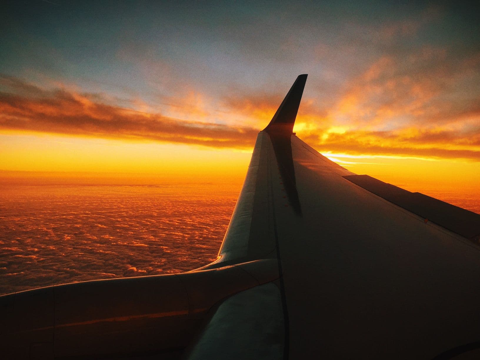 Airplane wing flying above clouds beneath a vivid orange sunset
