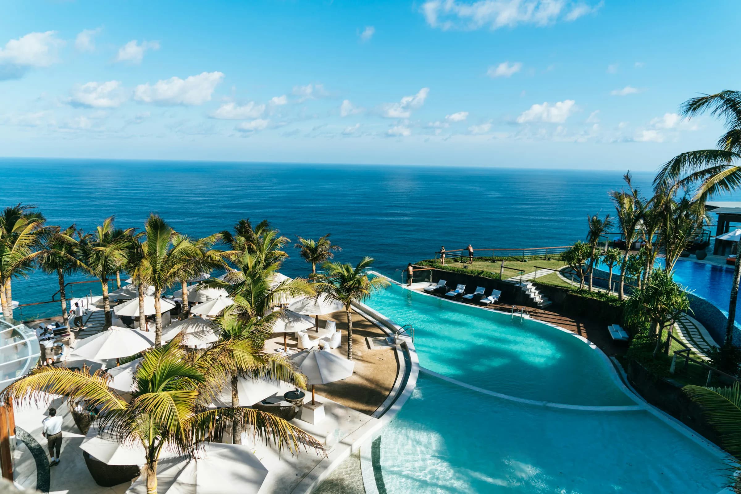Palm trees surrounding a turquoise oceanfront pool beneath a wide blue sky