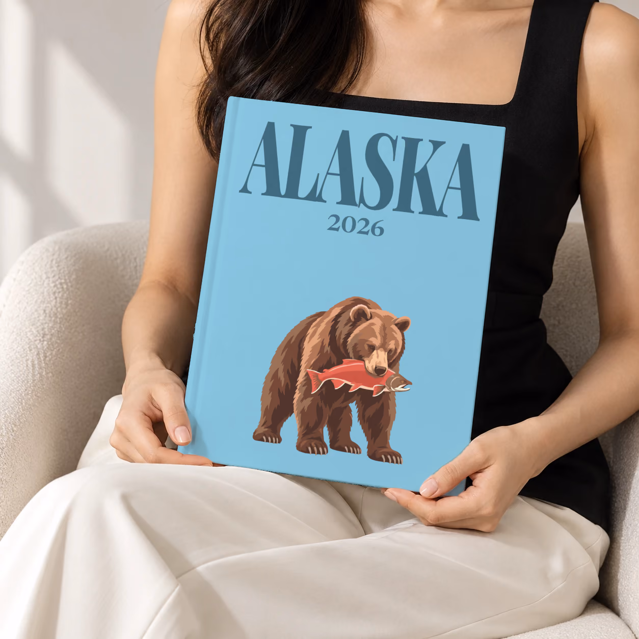 Alaska hardcover photo book with the illustrated cover of a grizzly bear catching a salmon on blue, held upright in two hands by a woman in a black top, sitting in a cream boucle armchair
