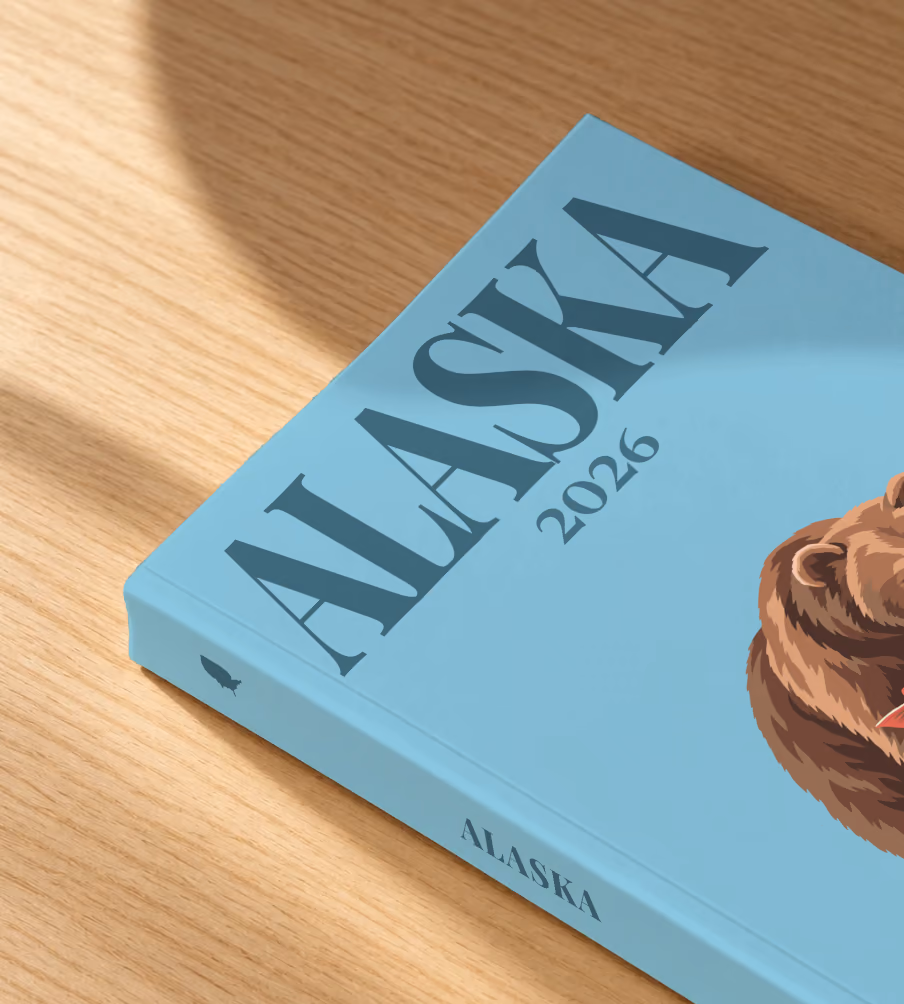 Alaska softcover photo book with the illustrated cover of a grizzly bear catching a salmon on blue, lying on an oak desk in dappled afternoon sunlight