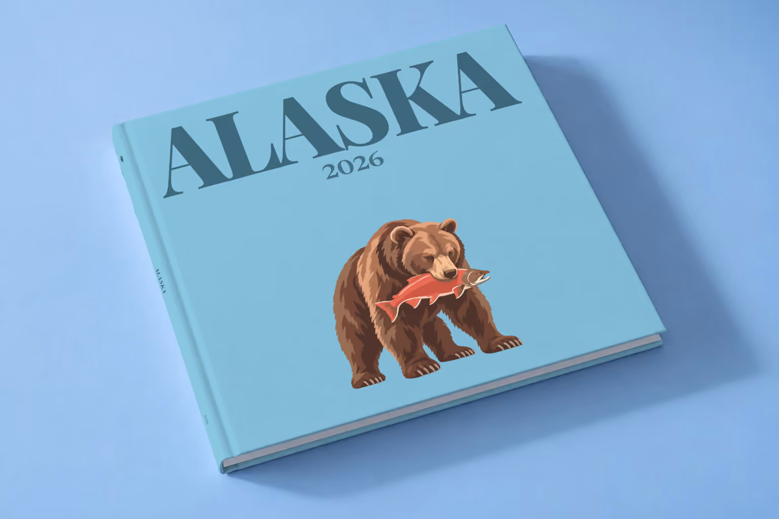 Alaska square hardcover photo book with the illustrated cover of a grizzly bear catching a salmon on blue, lying on a pale blue studio backdrop