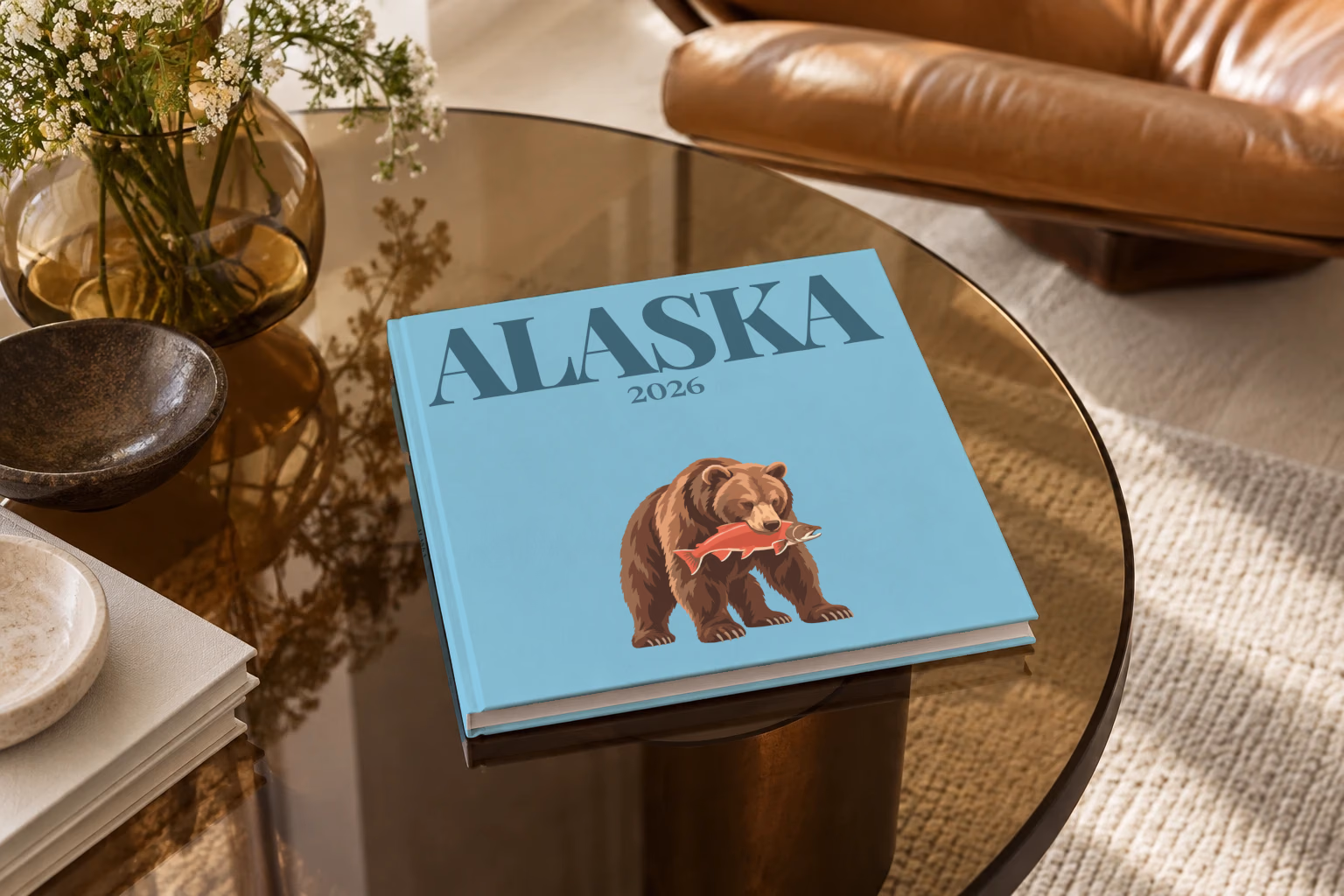 Alaska square hardcover photo book with the illustrated cover of a grizzly bear catching a salmon on blue, on a smoked glass coffee table beside a leather armchair in warm afternoon light