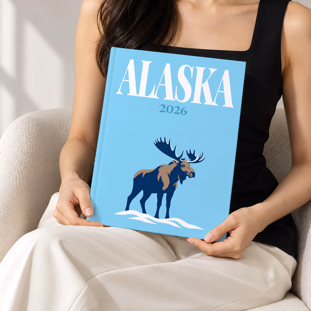 Alaska hardcover photo book with the illustrated cover of a navy moose in the snow on sky blue, held upright in two hands by a woman in a black top, sitting in a cream boucle armchair