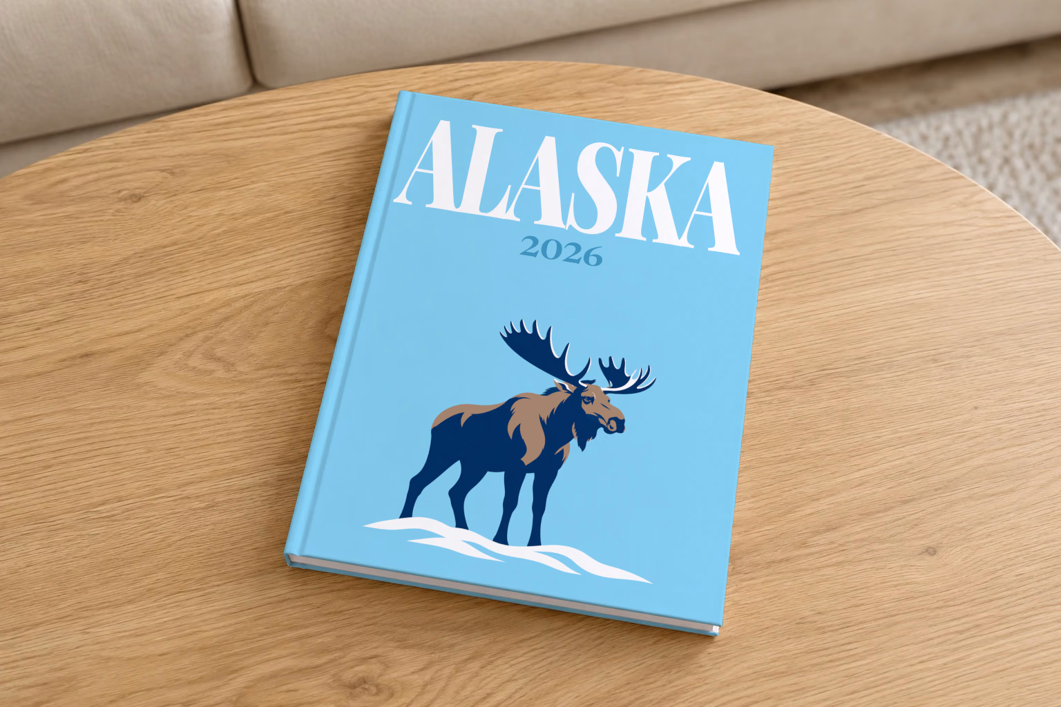 Alaska hardcover photo book with the illustrated cover of a navy moose in the snow on sky blue, on a round oak coffee table in a bright living room