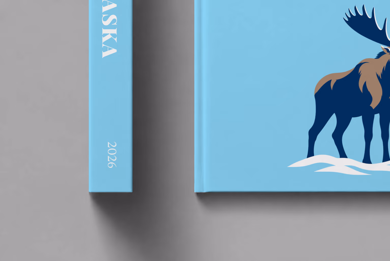 Alaska hardcover photo book with the illustrated cover of a navy moose in the snow on sky blue, shown close up beside its spine on a pale grey studio surface