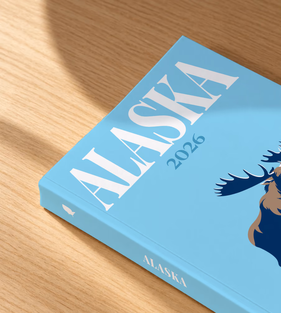 Alaska softcover photo book with the illustrated cover of a navy moose in the snow on sky blue, lying on an oak desk in dappled afternoon sunlight