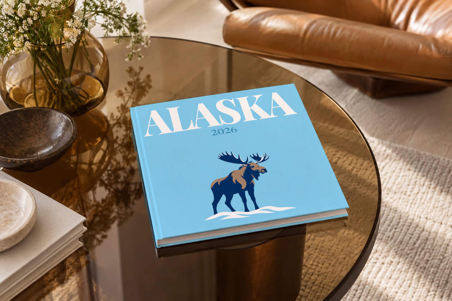Alaska square hardcover photo book with the illustrated cover of a navy moose in the snow on sky blue, on a smoked glass coffee table beside a leather armchair in warm afternoon light